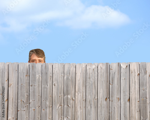 A goofy, happy man is being a peeping tom and nosy neighbour by stalking, watching and gawking over a wooden privacy fence