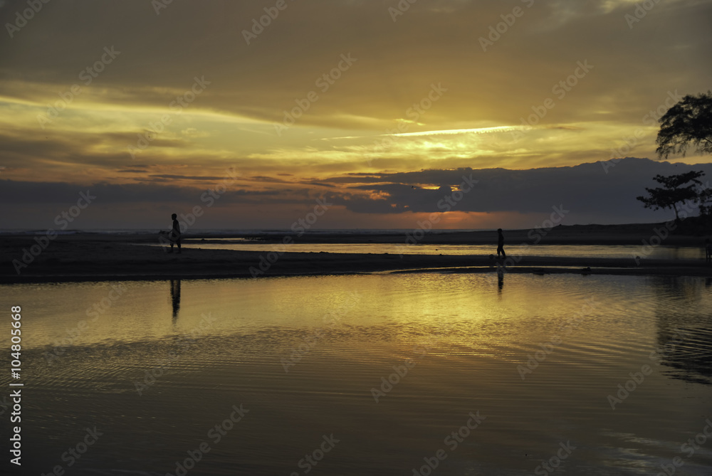 silhouette image of two boys walking inline at the shore with beautiful sunrise sunset background. soft clouds and reflection on the water