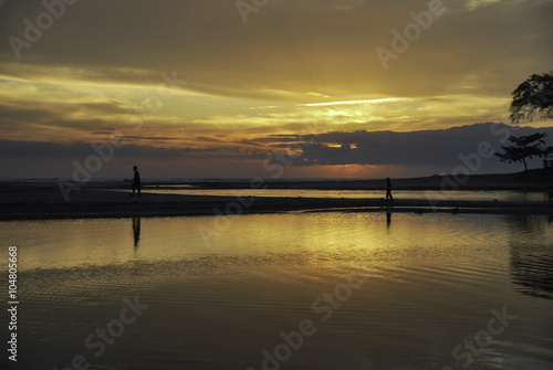 silhouette image of two boys walking inline at the shore with beautiful sunrise sunset background. soft clouds and reflection on the water