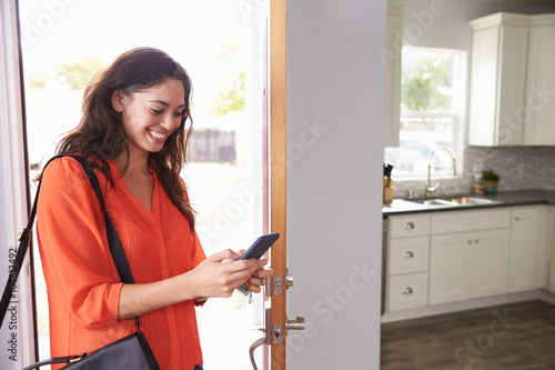 Tablou pe pânză Woman Checking Mobile Phone As She Opens Door Of Apartment