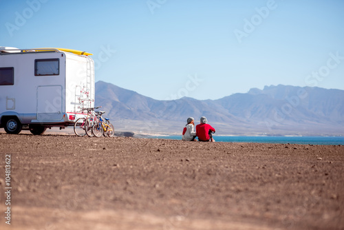 Young couple sitting near the camping trailer on the deserted seaside with mountains on background on Fuerteventura island in Spain