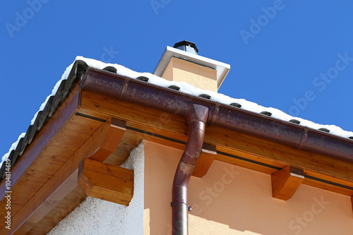 New roof top detail with ceramic tiles, chimney and copper water gutter with snow against blue skies