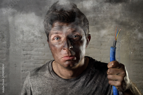 young man holding electrical cable smoking after electrical accident with dirty burnt face in funny sad expression