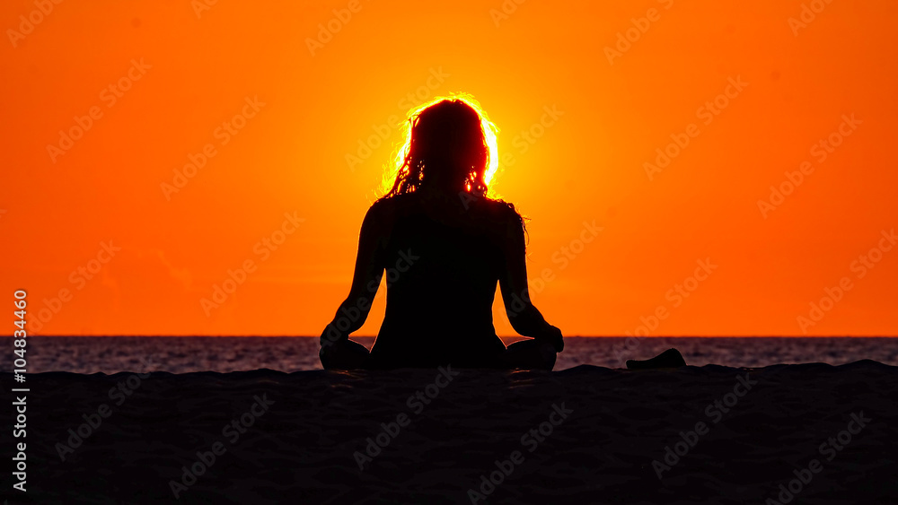 Silouette of meditating woman sitting in front of the red sinking sun on the beach