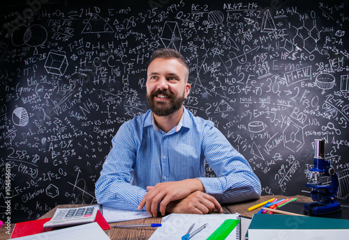 Happy teacher sitting at desk, school supplies, big blackboard