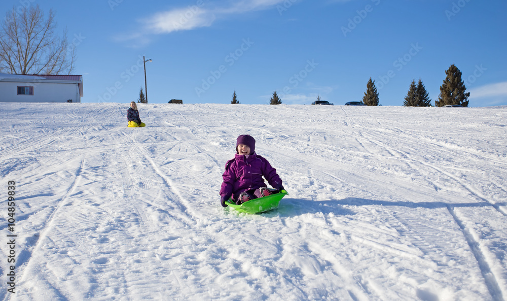 A little girl laughing going sledding downhill on a snow hill with a teenage girl sitting in the background