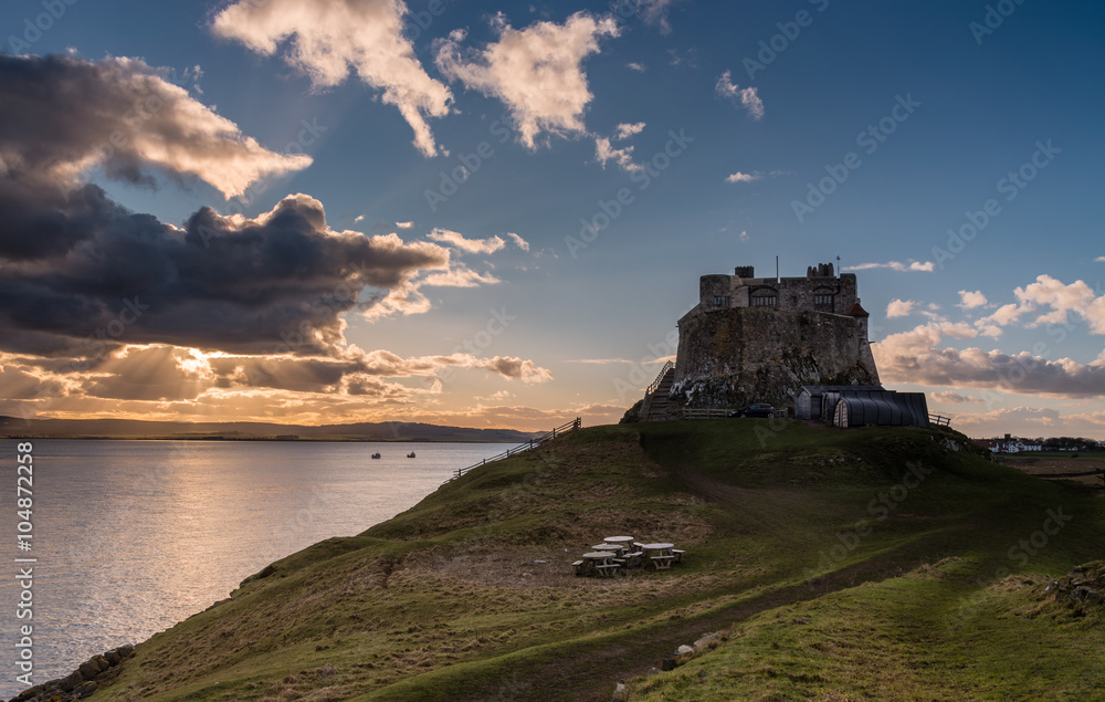 Lindisfarne Castle late afternoon with the suns orange rays bursting from behind clouds