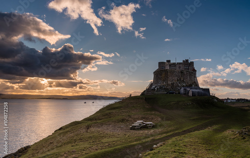 Lindisfarne Castle late afternoon with the suns orange rays bursting from behind clouds