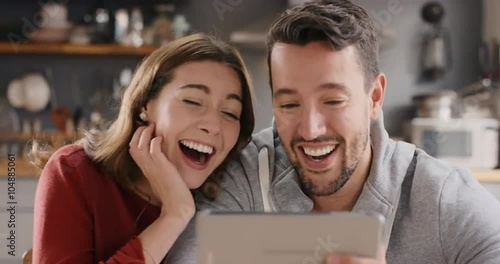 Attractive young couple at home laughing at funny internet joke online using digital tablet