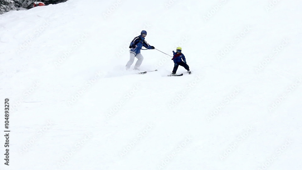 Young family, father and child, father holding the boy on a strap to control his speed, skiing in Austrian resort, wintertime Young family, father and child, father holding the boy on a strap to control his speed, skiing in Austrian resort, wintertime