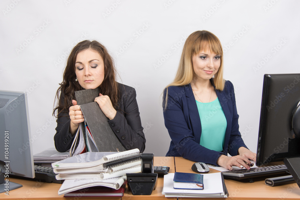 Female colleagues in the office, one asleep on a folder, the other happy working on a computer Female colleagues in the office, one asleep on a folder, the other happy working on a computer