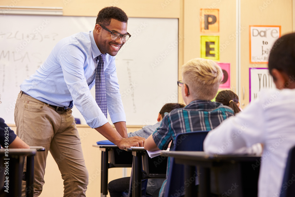 Smiling teacher leaning on elementary school pupil’s desk Smiling teacher leaning on elementary school pupil’s desk