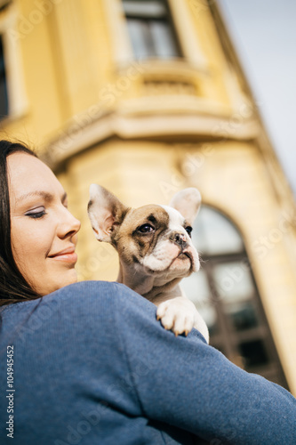 Young casually dressed woman holding her adorable French bulldog puppy. Close up shot with wide angle lens. Old, rustic building in background.