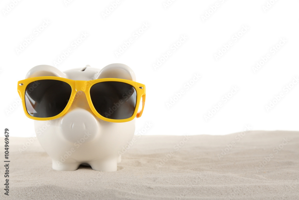 Piggy bank with sunglasses on a sand, isolated on white. Holiday money concept Piggy bank with sunglasses on a sand, isolated on white. Holiday money concept