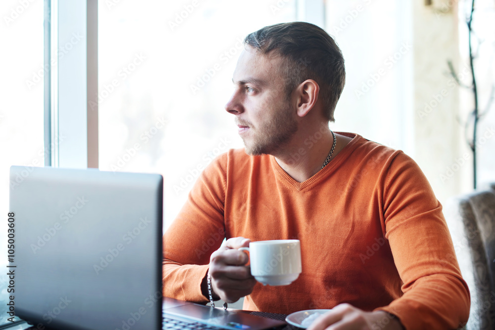 Handsome young man working on notebook, thinking, looking out the window, while enjoying coffee in cafe Handsome young man working on notebook, thinking, looking out the window, while enjoying coffee in cafe