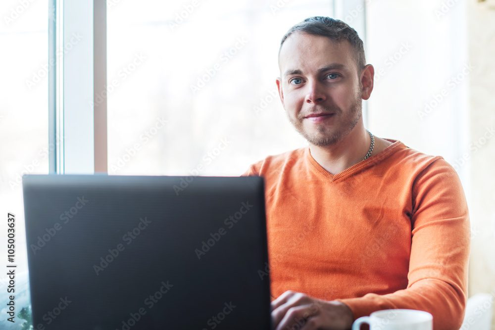 Fototapeta premium Handsome young man working on notebook, smiling, looking at camera, while enjoying coffee in cafe