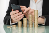 Businesswoman Using Smartphone With Stacked Coins On Desk