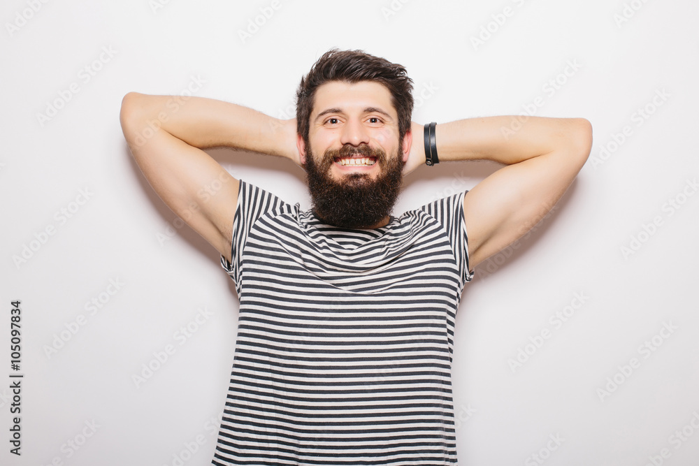 Handsome young man with beard holding hands behind head and smiling while standing against white background Handsome young man with beard holding hands behind head and smiling while standing against white background