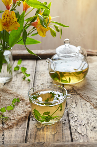 Melissa tea in glass cup, transparent teapot with leaves and yellow flowers. Healthy natural organic beverage on vintage wooden table background.