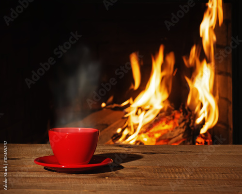 Fototapeta Red cup of tea or coffee near fireplace on wooden table.