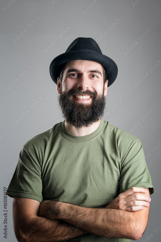 Bearded hipster wearing hat with upper lips piercing friendly laughing at camera.  Headshot portrait over gray studio background with vignette.