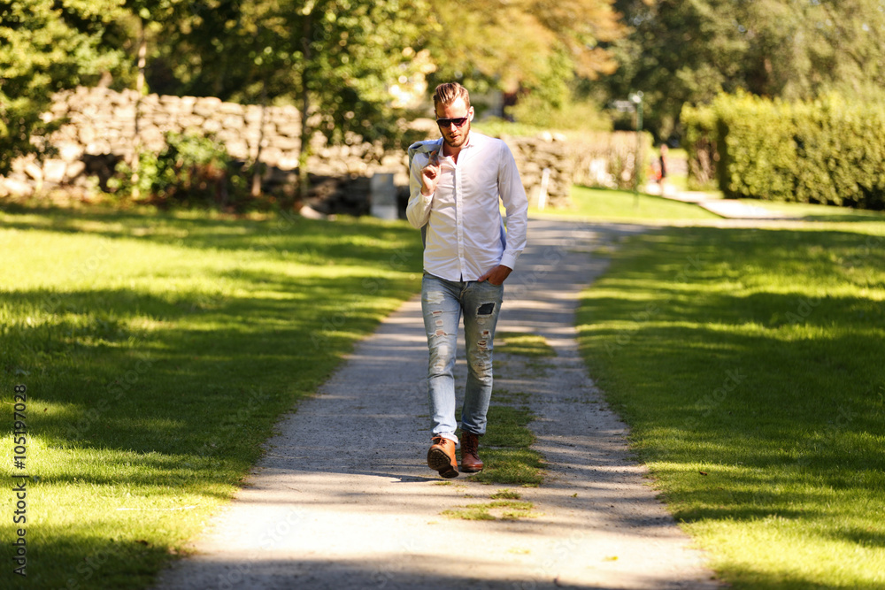 An attractive fashion model walking along a path outdoors on a sunny summer day, holding his light blue jacket over his shoulder. An attractive fashion model walking along a path outdoors on a sunny summer day, holding his light blue jacket over his shoulder.