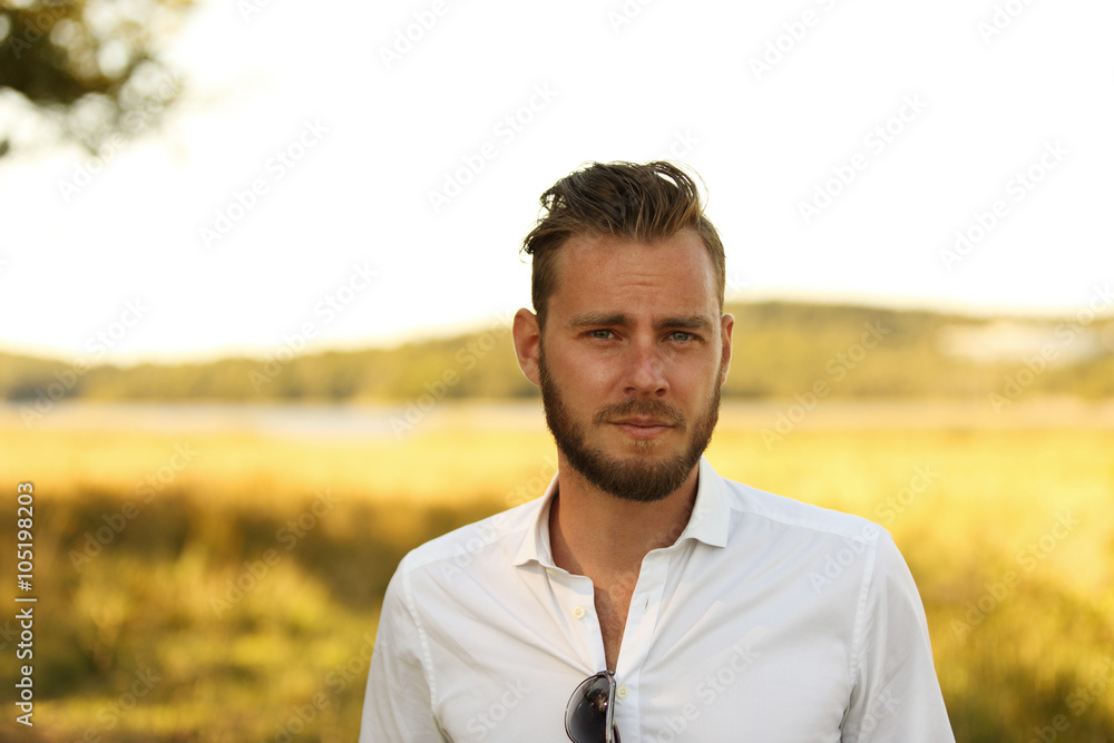 Man with a beard wearing a white shirt standing in front of a big yellow field with the sun behind, thinking and lonely.