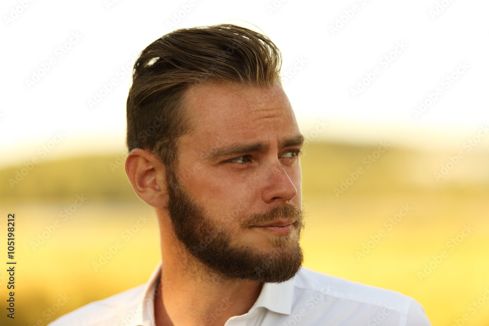 Man with a beard wearing a white shirt standing in front of a big yellow field with the sun behind, thinking and lonely. Man with a beard wearing a white shirt standing in front of a big yellow field with the sun behind, thinking and lonely.