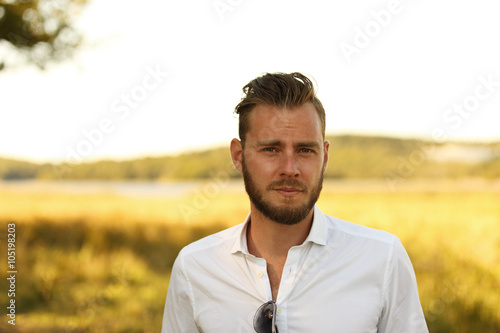 Man with a beard wearing a white shirt standing in front of a big yellow field with the sun behind, thinking and lonely.