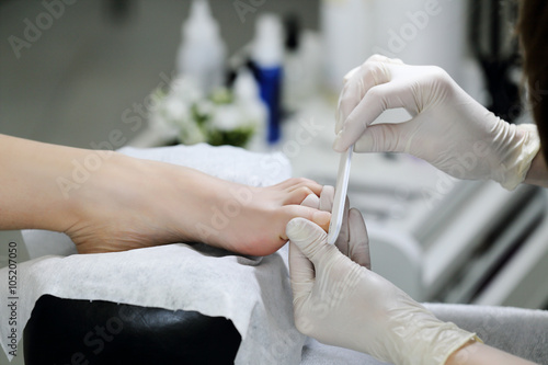 The big toe is doing pedicure nail file in salon. Manicurist in white mittens works with the client.