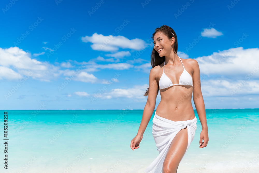 Sexy bikini woman relaxing on beach with slim stomach wearing white triangle top and pareo cover up beachwear - weight loss or epilation concept.