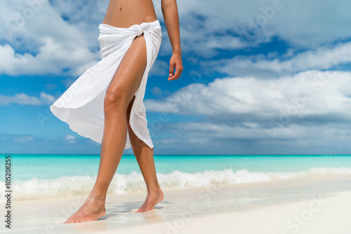 White pareo woman legs walking on tropical beach vacation. Closeup of barefoot female young adult lower body relaxing in ocean water on summer holiday travel wearing cover-up beachwear.  photo