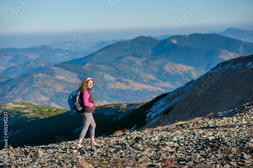 Young happy woman backpacker is walking on mountain ridge with beautiful mountains on background. Sunny autumn day.