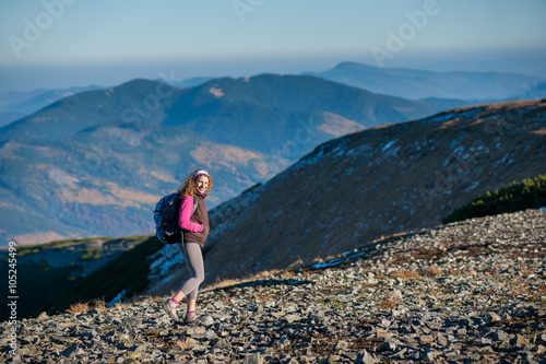 Young happy woman backpacker is walking on mountain ridge with beautiful mountains on background with big backpack on, looking to the camera. Sunny autumn day.