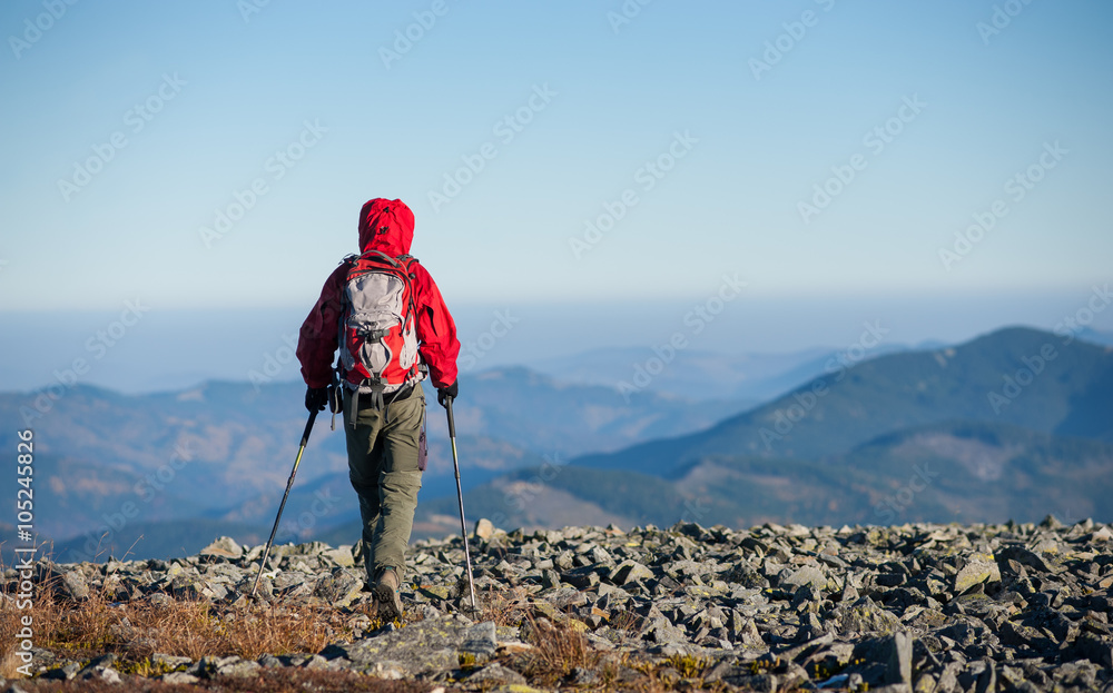 Male sportsman backpaker walking on the rocky top of the mountain with beautiful mountains on background. Man is wearing red jacket and has trekking sticks and backpack on. Rear view.