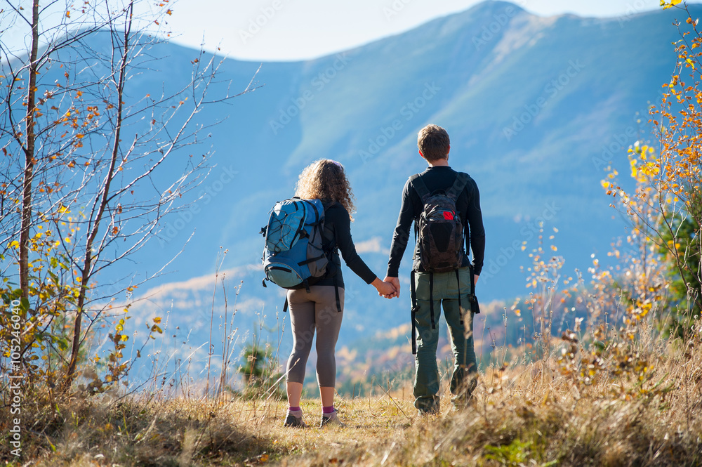 Rear view of young backpackers couple looking into distance enjoying the overlook view and holding hands on a warm sunny autumn day in the mountains. Mountain on the background. Warm fall time.