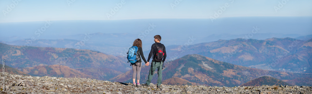 Rear view of young couple hikers with backpacks standing on the ridge of the mountain, enjoying the panorama view of beautiful open overlook on the mountains. Couple is holding hands. Wide angle view