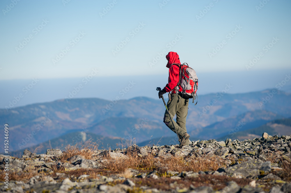Male sportsman backpaker walking on the rocky top of the mountain with beautiful mountains on background. Man is wearing red jacket and has trekking sticks and backpack on.