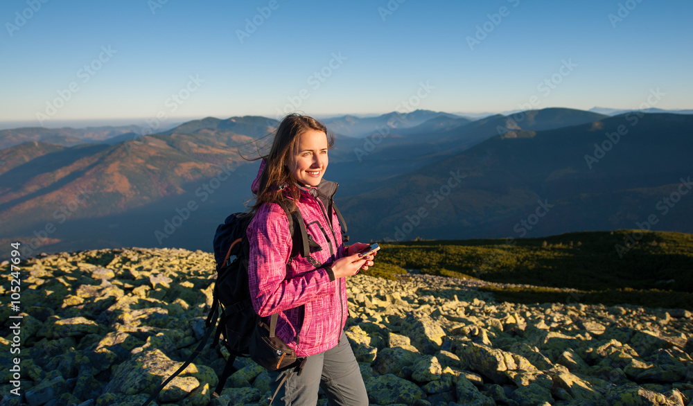 Portrait of young cute woman backpacker standing on rocky top of the mountain, looking at the sun and holding cell phone. Side view. Ecotourism and healthy lifestyle concept. Sunny fall evening.