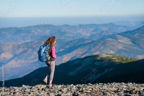 Young woman tourist is walking on mountain plato with big backpack on. Beautiful mountains on background. Sunny autumn day.
