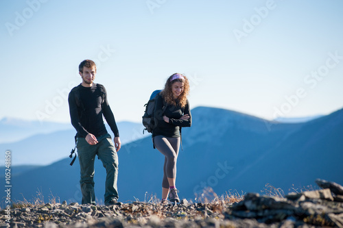 Portrait of male and female hikers walking up in the mountains. Front view with beautiful mountains on the background. Girl is smiling and looking down, guy is looking into the camera.