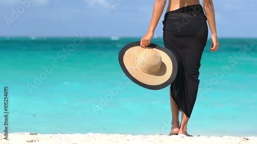 Lady on beach in black sarong standing on shore looking at ocean. Sexy female is enjoying wind on beach. Woman is holding sunhat during summer vacation by turquoise sea in the Caribbean.