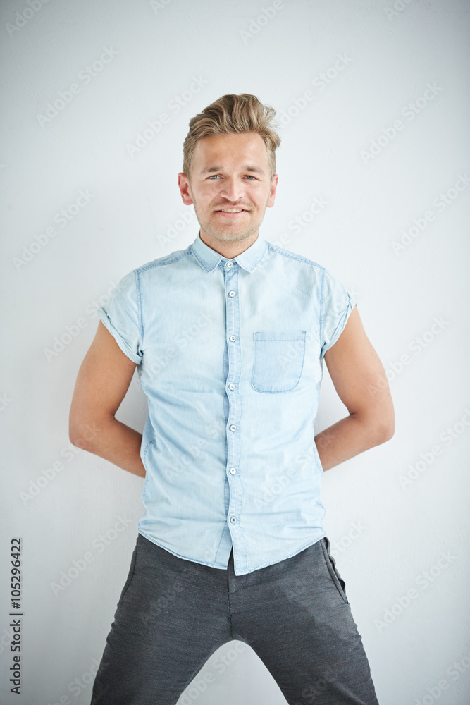 Brutal man with a small beard is leaning on the wall , wearing a blue short-sleeved shirt and gray pants , hands are behind his back Brutal man with a small beard is leaning on the wall , wearing a blue short-sleeved shirt and gray pants , hands are behind his back