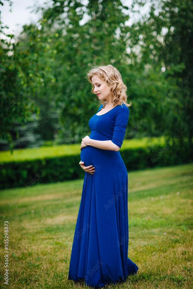 Pretty young pregnant woman in blue dress with long blond curly hair holding her belly and looking at it in summer park on rainy day. Pregnancy and femininity concept. Waterdrops on blue dress Pretty young pregnant woman in blue dress with long blond curly hair holding her belly and looking at it in summer park on rainy day. Pregnancy and femininity concept. Waterdrops on blue dress