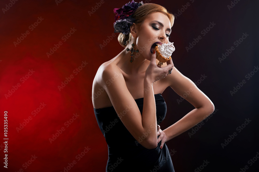 Fashionable eating. Studio shot of a gorgeous model wearing black dress and flowers in her hair posing biting a cupcake Fashionable eating. Studio shot of a gorgeous model wearing black dress and flowers in her hair posing biting a cupcake