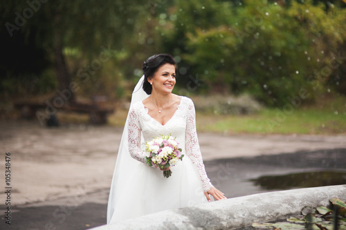 Beautiful brunette bride in elegant white dress holding bouquet posing neat trees 