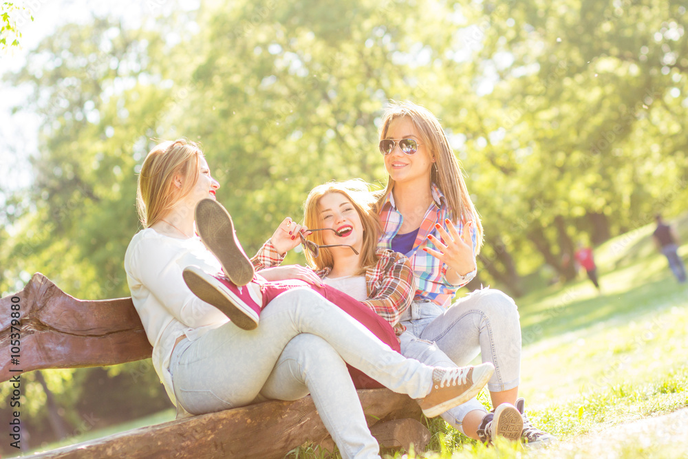 Group of three teenage girls sitting and chatting on bench in park.
