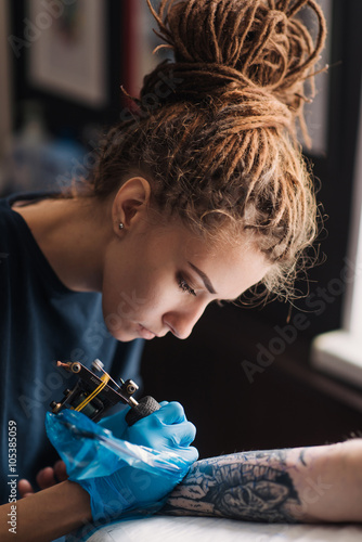 Tattoo girl with dreadlocks on his head in the stuffing sleeve tattoo in black and white. Master works in a professional salon on a white mat and in sterile blue gloves.