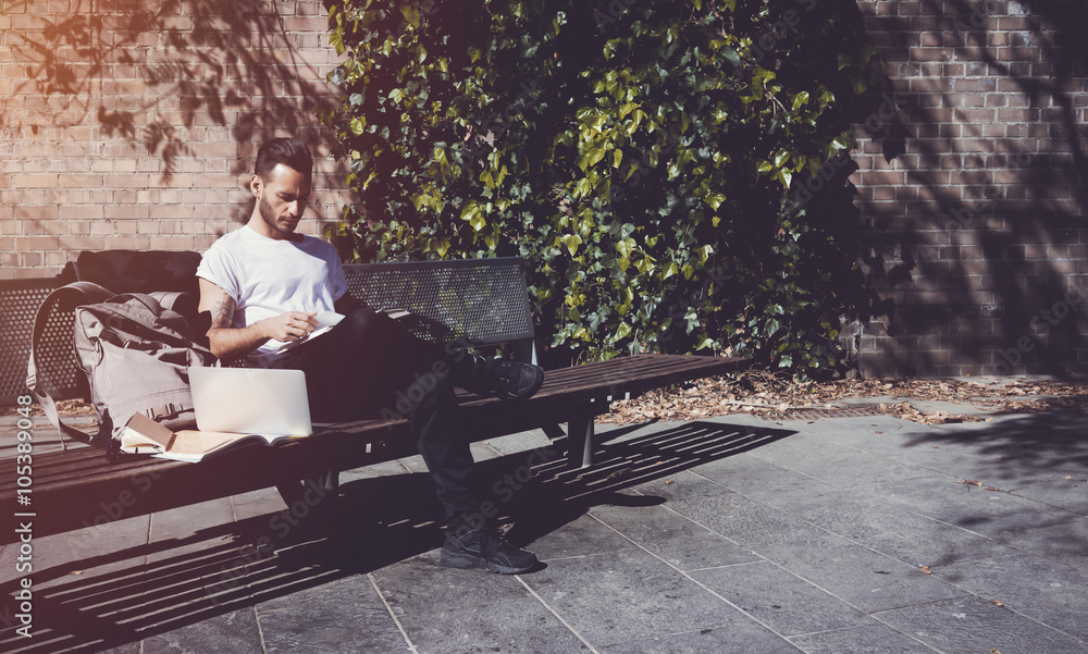 Young man wearing white tshirt sitting city park and reading book. Studying at the University, working new project. Books, laptop, backpack bench. Horizontal, film effect Young man wearing white tshirt sitting city park and reading book. Studying at the University, working new project. Books, laptop, backpack bench. Horizontal, film effect