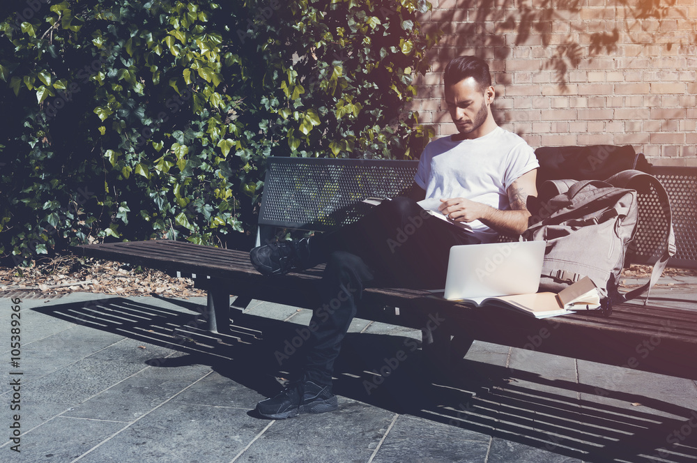 Fototapeta premium Photo bearded man wearing white tshirt sitting city park bench and reading book. Studying at the University, working new project. Books, laptop, backpack bench. Horizontal mockup